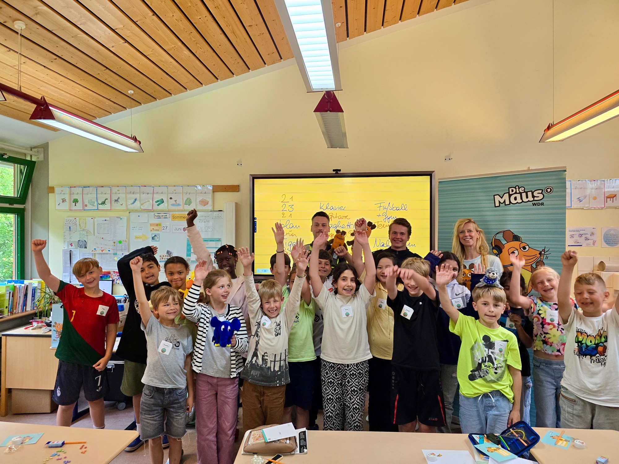 Das Bild zeigt eine fröhliche Grundschulklasse, die mit erhobenen Armen in einem Klassenzimmer steht. In der Mitte stehen zwei Erwachsene und eine Frau vor einem „Die Maus“-Banner. Im Hintergrund ist ein großer Bildschirm mit einer digitalen Tafel zu sehen. Die Stimmung ist ausgelassen – es handelt sich um eine Schulaktion der „Maus-Klasse“.