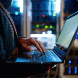 Ein Mann steht mit Laptop in einem Serverraum. Schemenhafte Darstellung