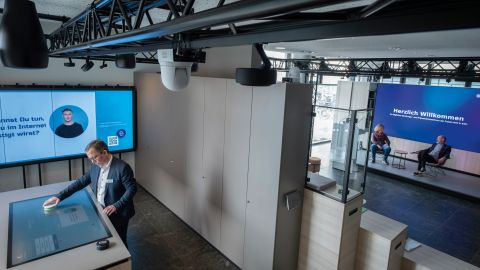 The concept store is divided into two separate areas. In the left-hand area is the table with touch function. Chief Inspector Thomas Jansen is operating it. In the right-hand area, a large screen can be seen on the wall. In front of it, two people are sitting in a seating area on a slightly raised platform.