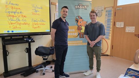 Das Bild zeigt zwei junge Männer in einem Klassenzimmer, die vor einem „Sendung mit der Maus“-Banner stehen und lächeln. Links ist ein großer Bildschirm mit einer digitalen Tafel zu sehen, auf der Begriffe wie „Klavier“, „Fußball“ und „Reporter“ stehen. Der Raum wirkt kindgerecht – es ist eine Schulaktion.